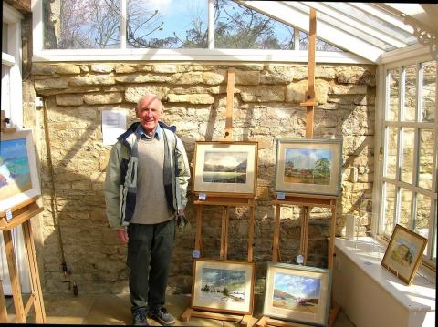 Derek stands to the left of an arrangement of his paintings, all displayed on easels