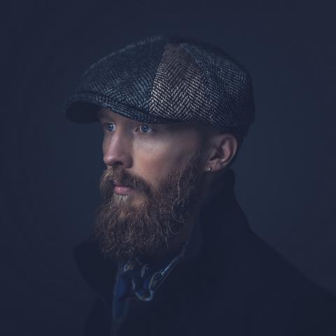 A young man with a large ginger beard looks off into the distance. He has blue eyes. He wars a blue coat with the collar turned up to cover his neck and a grey tweed flat cap on his head. The portrait shows the man from the chest up.