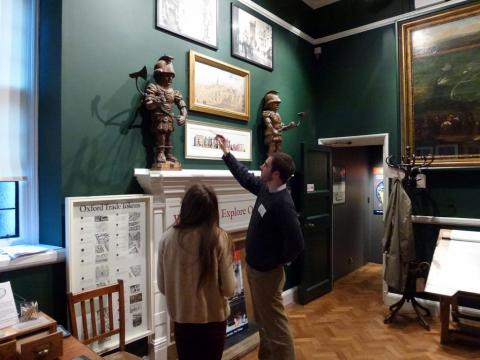 A volunteer stands in the museum next to a visitor, he points to one of the 'quarter boy. statues on the shelf, figures that once were part of the mechanism that rang the bells at Carfax Tower