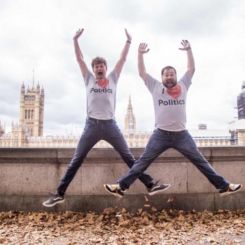 2 men in I heart politics t-shirts are jumping against a backdrop of the Houses of Parliament