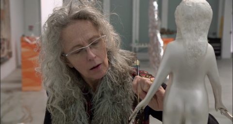Kiki Smith painting a silver sculpture of a female figure with a paintbrush.