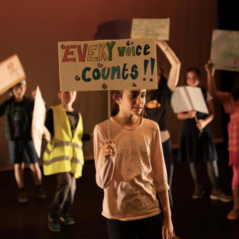 Group of young people on stage - one holds a placard saying 'Every Voice Counts'