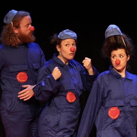 Three clowns stand looking confused - they are dressed in blue boiler suits and have red noses on