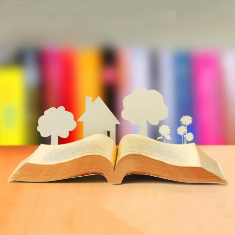 An open book lies on a table. Paper cutouts of a house and trees are popping out of it.