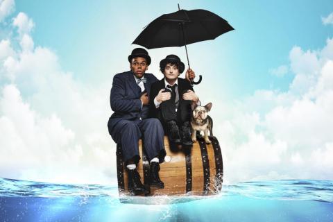Actors dressed as Stan Laurel and Charlie Chaplin sit with a dog atop a barrel floating on water. Laurel holds a black umbrella above Chaplin 
