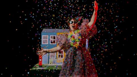 woman in a multi-colour dress throwing confetti in the air, blue doll house behind her