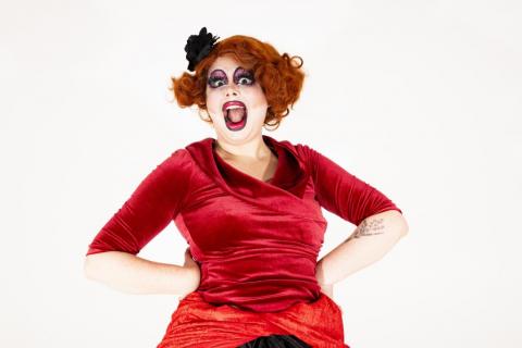 Ginger, a white drag queen with heavily clown-like makeup, including massively overdrawn red lips, bold purple eyeshadow, and big fake eyelashes. They have a short curly ginger wig, with a black rose worn on the right side of their head. They are photographed from the waist up against a white background, wearing a red velvet blouse with sleeves down to the elbow, and have a floral tattoo on their left forearm. They are posing with their hands on their hips and a wide open smile in a look of joyous surpris