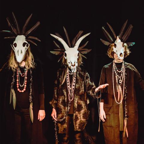 3 women in paper skull masks and ornamental robes stand ready to sing, one of them carries a red bass guitar.