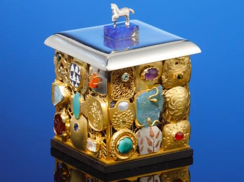 Blue background. In the centre is the artwork with its reflection below. A small silver horse sits on the top surface. Top surface is a square thick shelf of silver metal, resting on the decorated cube below. Each face of the cube has 9 smaller panels arrange in three rows and thee columns. Each panel is different, but they are all mostly gold.