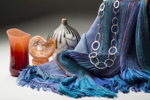Arrangement of craft items. White surface, dark grey and black background. On the right a blue textile has been draped over and covering a  box so that it cascades down the right of the image and most of the foreground. A silver chain of large rings is draped over the textile. On the left are three ceramic and glass vases.