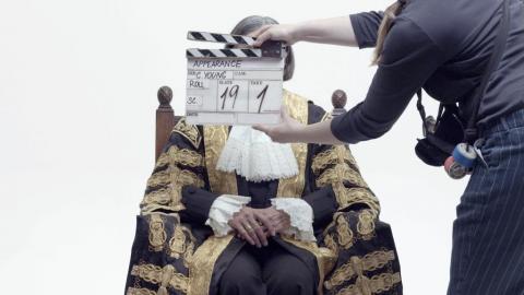 A film clapperboard being held in front of a person in an ornate gold and black costume, who is sitting with their hands in front of them against a white background.