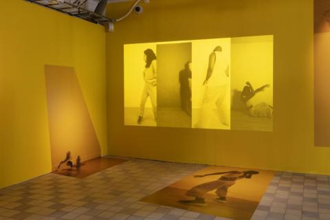 An art piece by Harold Offeh. A yellow glowing room with images projected on to the wall and floor. Pools of warm coloured light filter through.