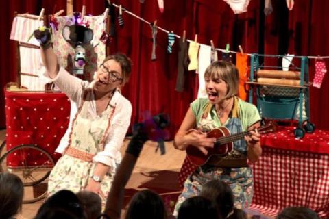 Two women sit in front of a crowd of children holding hand made sock puppets. One also plays a ukulele.