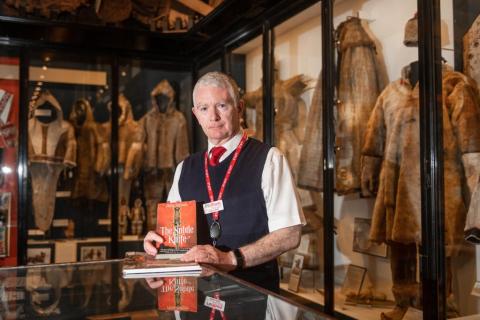 Male member of museum staff holding book in front of display cases full of Arctic clothing