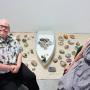 A man and a woman sitting on chairs with a boat and stones art installation 
